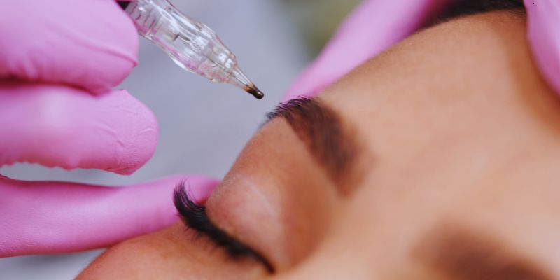 Close-up of a woman receiving a microblading touch up procedure with pink-gloved hands carefully shaping eyebrows using a precision tool.