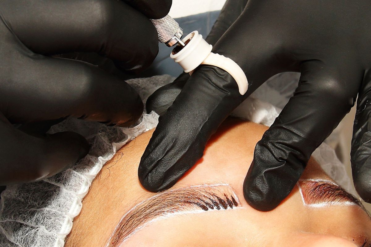 Professional performing a microblading touch up on eyebrows with black gloves and specialized pigment tool for defining brow strokes