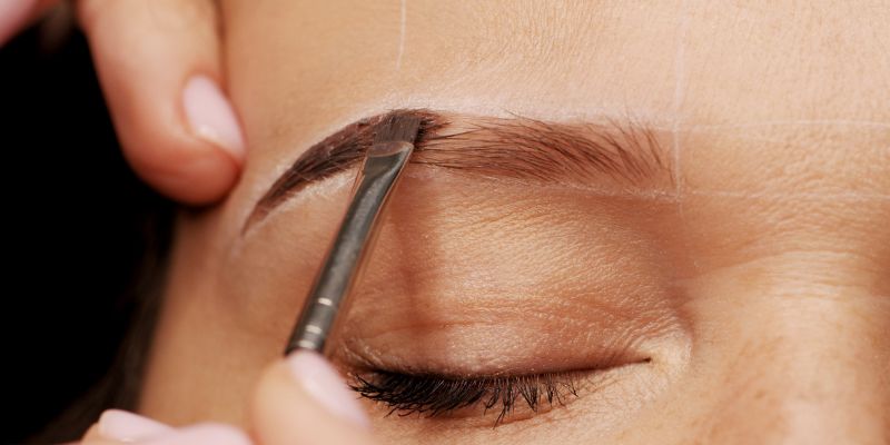 Close-up of a professional applying dark tint with a small brush during eyebrow tinting procedure, shaping and filling in the brows on a client with closed eyes.