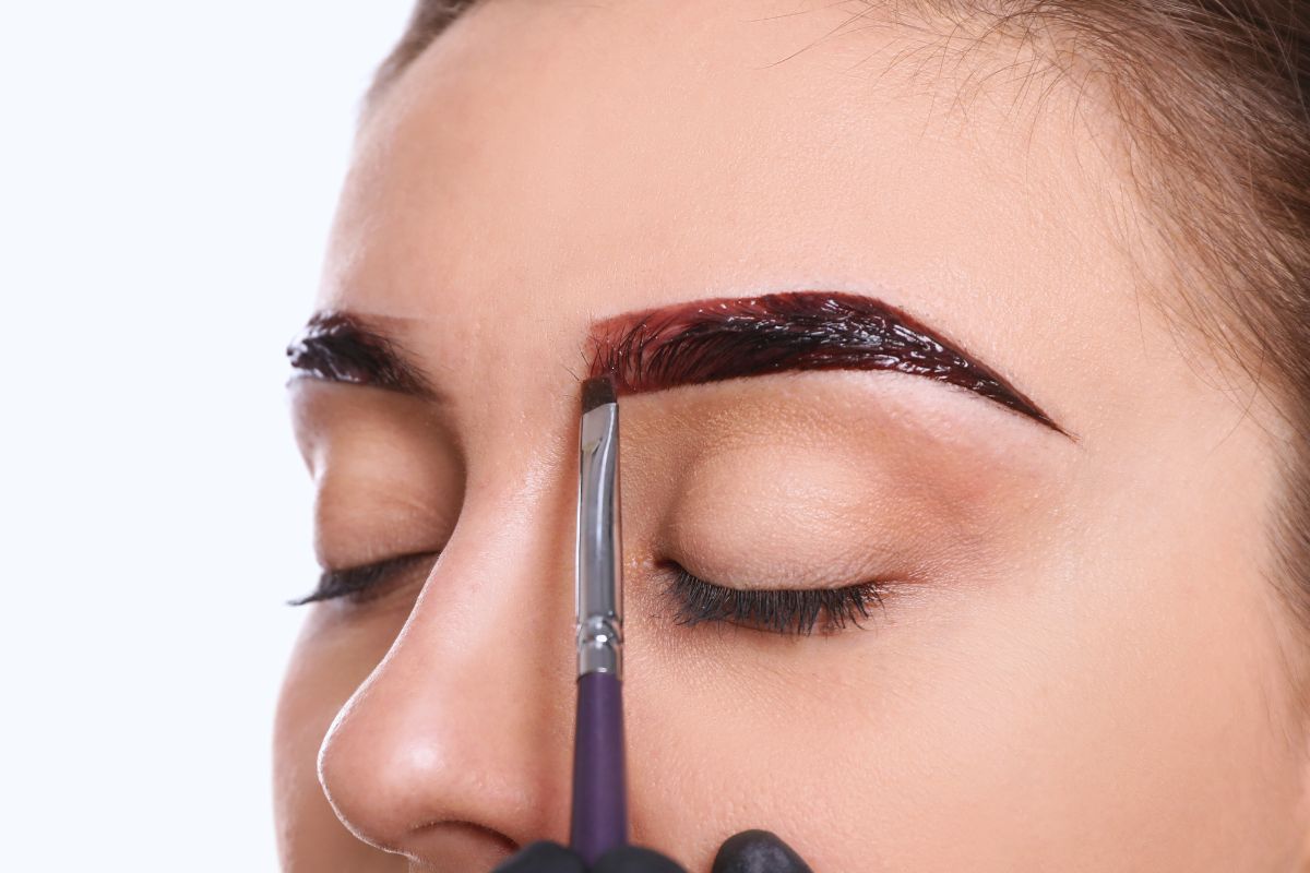 Beautician using a precise angled brush to apply rich dark tint to perfectly shaped eyebrows in an eyebrow tinting service, achieving bold defined results on a client with relaxed expression.