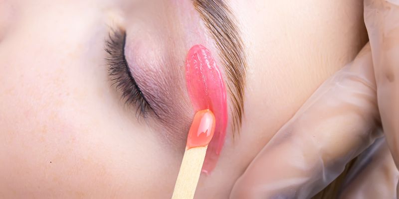 How often to get eyebrows waxed: professional esthetician applying warm golden wax precisely to shape and remove unwanted eyebrow hair during a brow waxing session