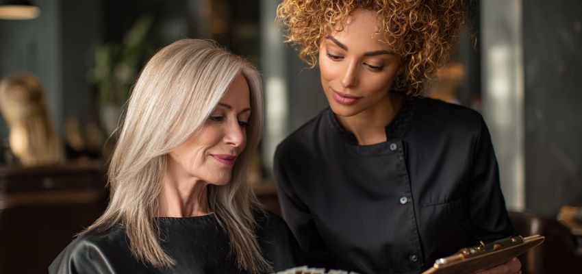A soft warm ash blonde long hairstyle on a mature woman is featured during a salon consultation in the warm blonde vs cool blonde comparison.